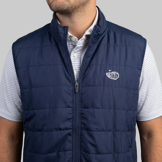 A man wearing a navy blue quilted puffer vest over a white and blue striped button-down shirt. The vest features a stand-up collar, front zipper, and a small white embroidered wave logo on the left chest.