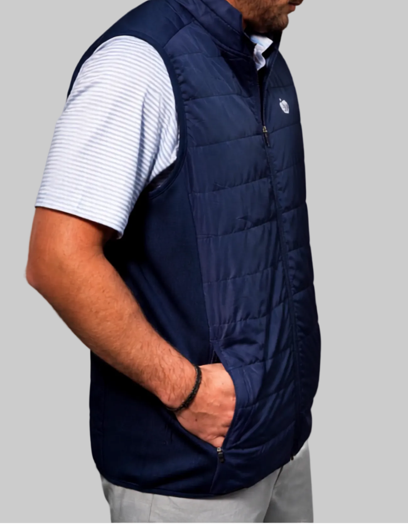 A man in profile wearing a navy blue quilted zip-up vest over a white and blue striped polo shirt, a white baseball cap, and light gray pants, photographed against a neutral gray studio background.
