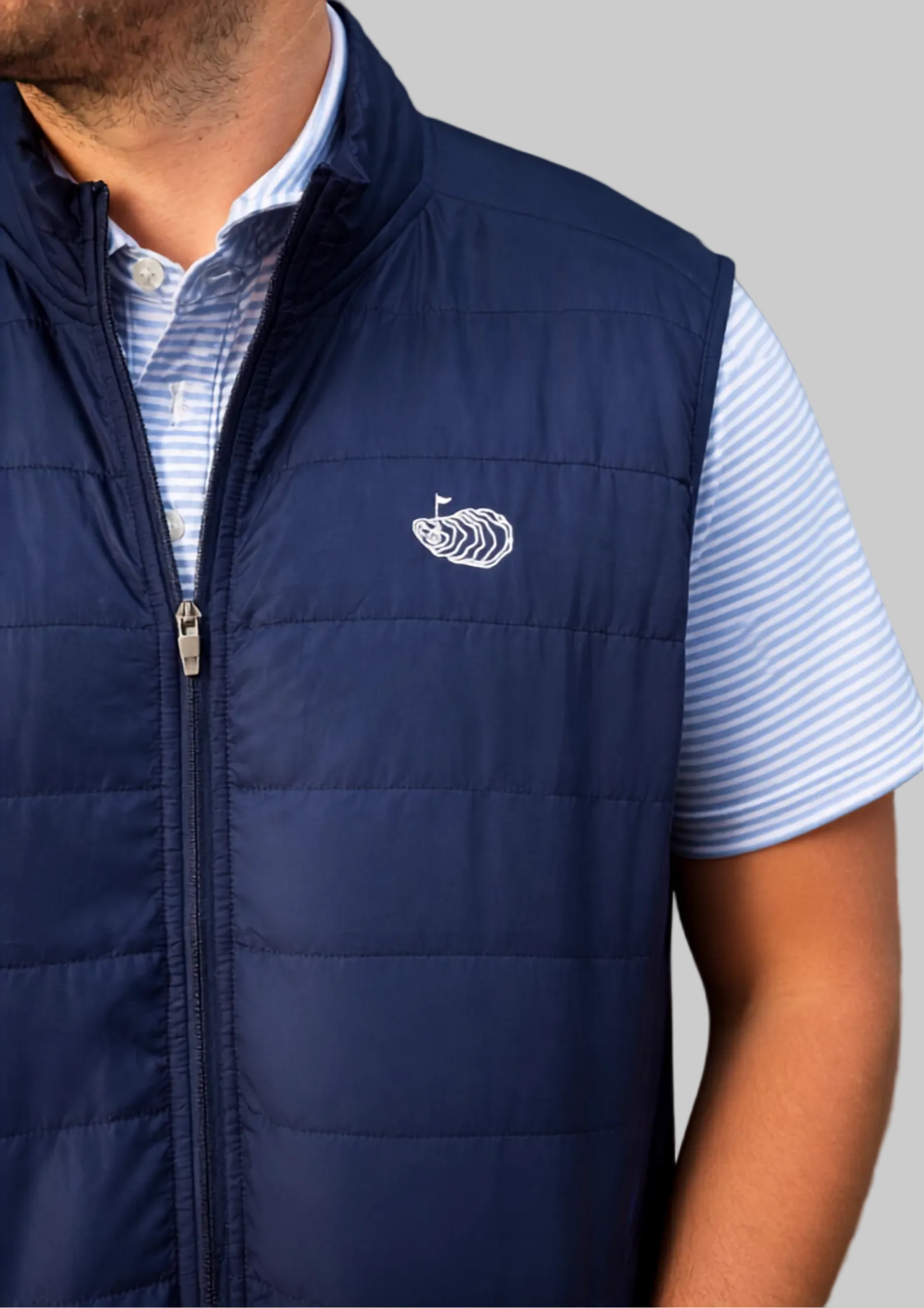 A close-up of a man wearing a navy blue quilted vest over a light blue and white striped polo shirt. The vest features a white embroidered boat logo on the left chest, characteristic of the brand Vessel.